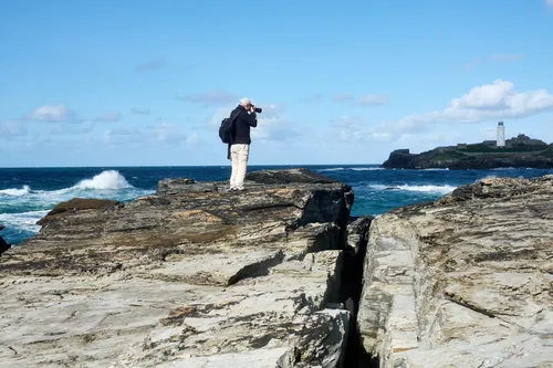Godrevy photographer This landscape photograph captures a coastal scene in Cornwall during a late morning in early autumn. The main subject, a person identified as Michael Weinbren, stands on the rugged rocks of the Godrevy coast, taking photographs of the scenery. In the background, the well-known Godrevy Lighthouse is visible on the headland, overlooking the Atlantic Ocean. The image shows waves crashing against the rocks under a partly cloudy sky, with the presence of people adding scale and context to this iconic Cornwall location.